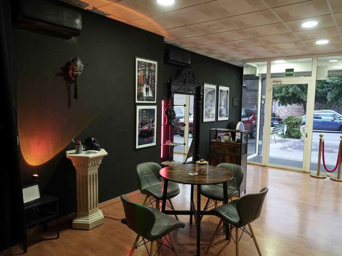 Zona de espera de Dome Shop Tattoo en Terrassa: paredes negras, mesa redonda con sillas, una columna clásica, pósters de cine y la entrada acristalada a la calle