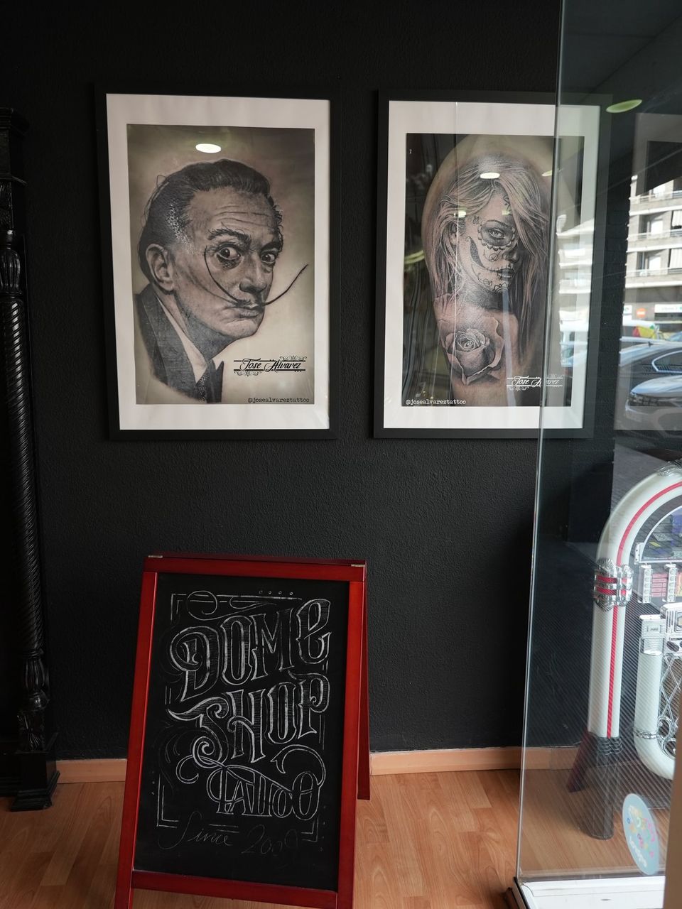 Pizarra con el logotipo «Dome Shop Tattoo since 2009» dibujado a tiza, bajo dos fotos enmarcadas de retratos realistas tatuados por Jose Alvarez