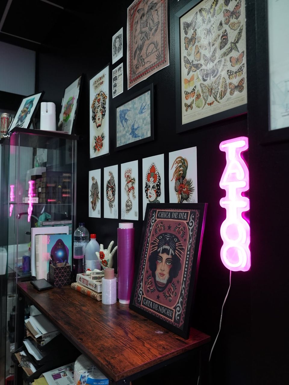 Rincón de trabajo con láminas de tatuaje tradicional, un cartel de neón rosa «Tattoo» y una vitrina con material