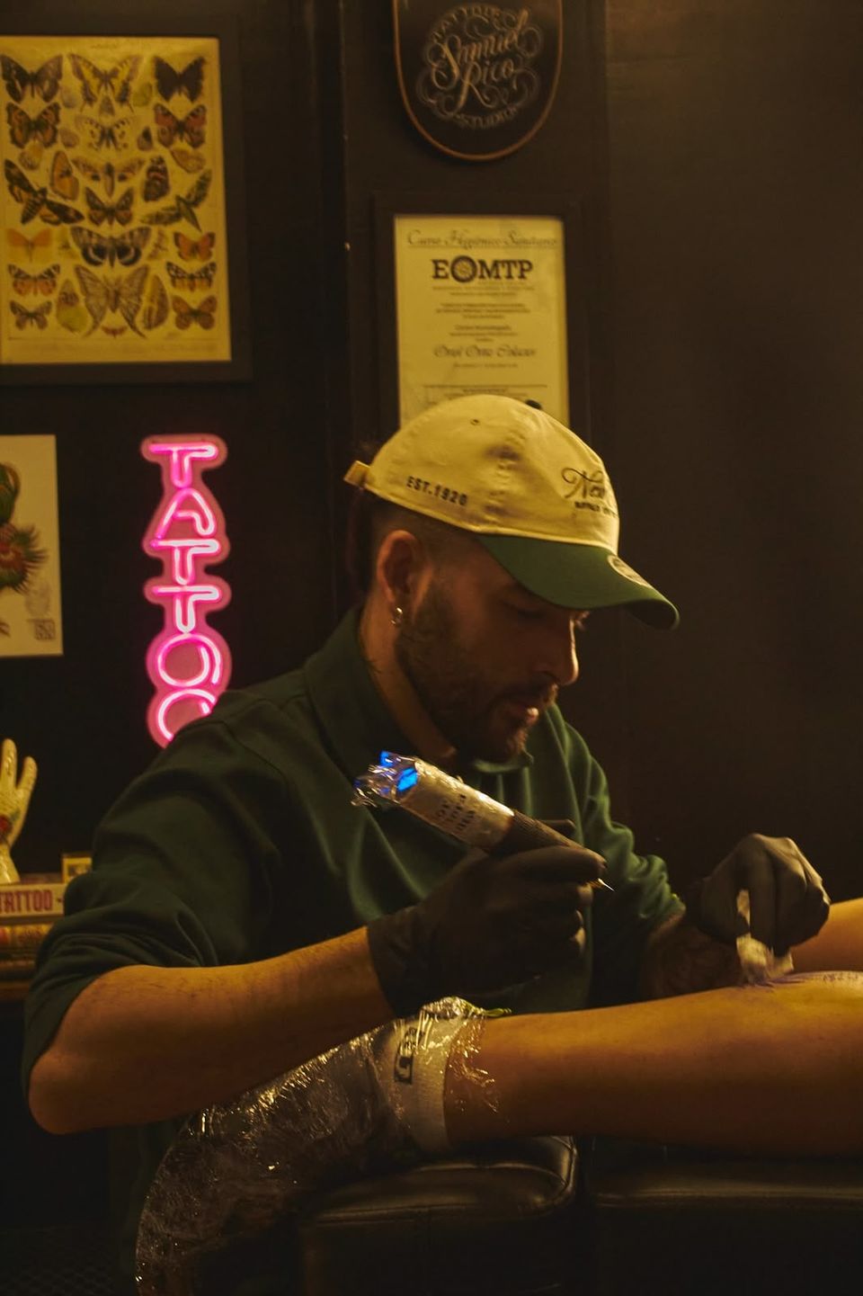 Uri Ortiz tatuando una pierna en Dome Shop Tattoo, con un neón rosa de «Tattoo» al fondo