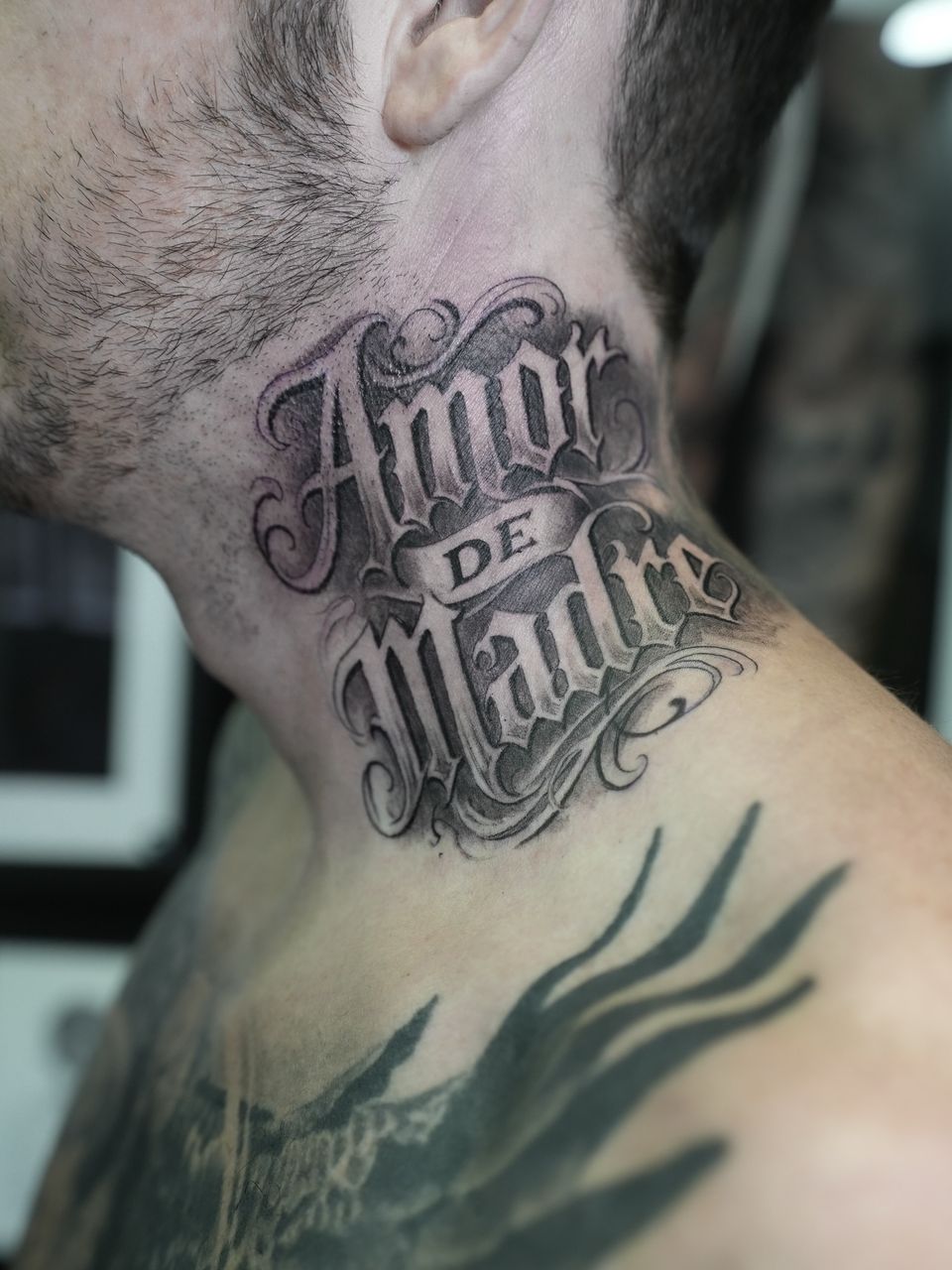 Tatuaje de letras en el lateral del cuello: «Amor de Madre» en letra gótica ornamental con sombreado, en negro y grises