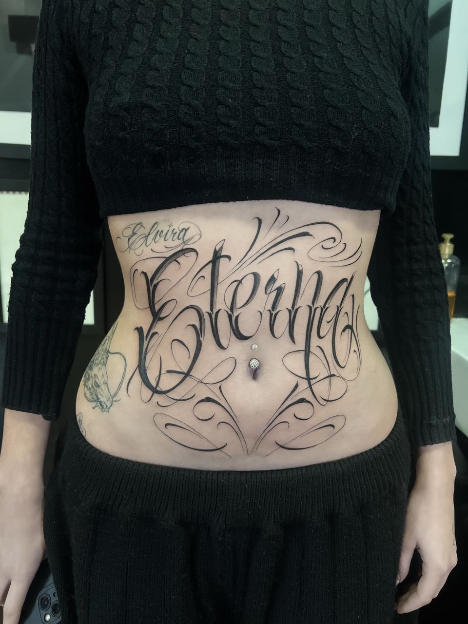Tatuaje de letras en el abdomen: la palabra «Eterna» en caligrafía con grandes adornos y un nombre más pequeño encima, en negro