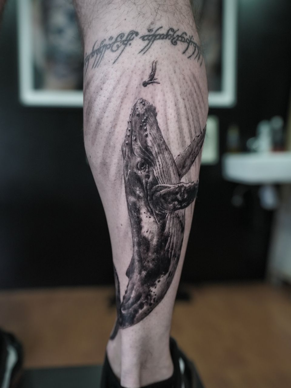 Tatuaje realista en el gemelo: ballena jorobada ascendiendo hacia un pequeño buceador entre rayos de luz, en negro y grises
