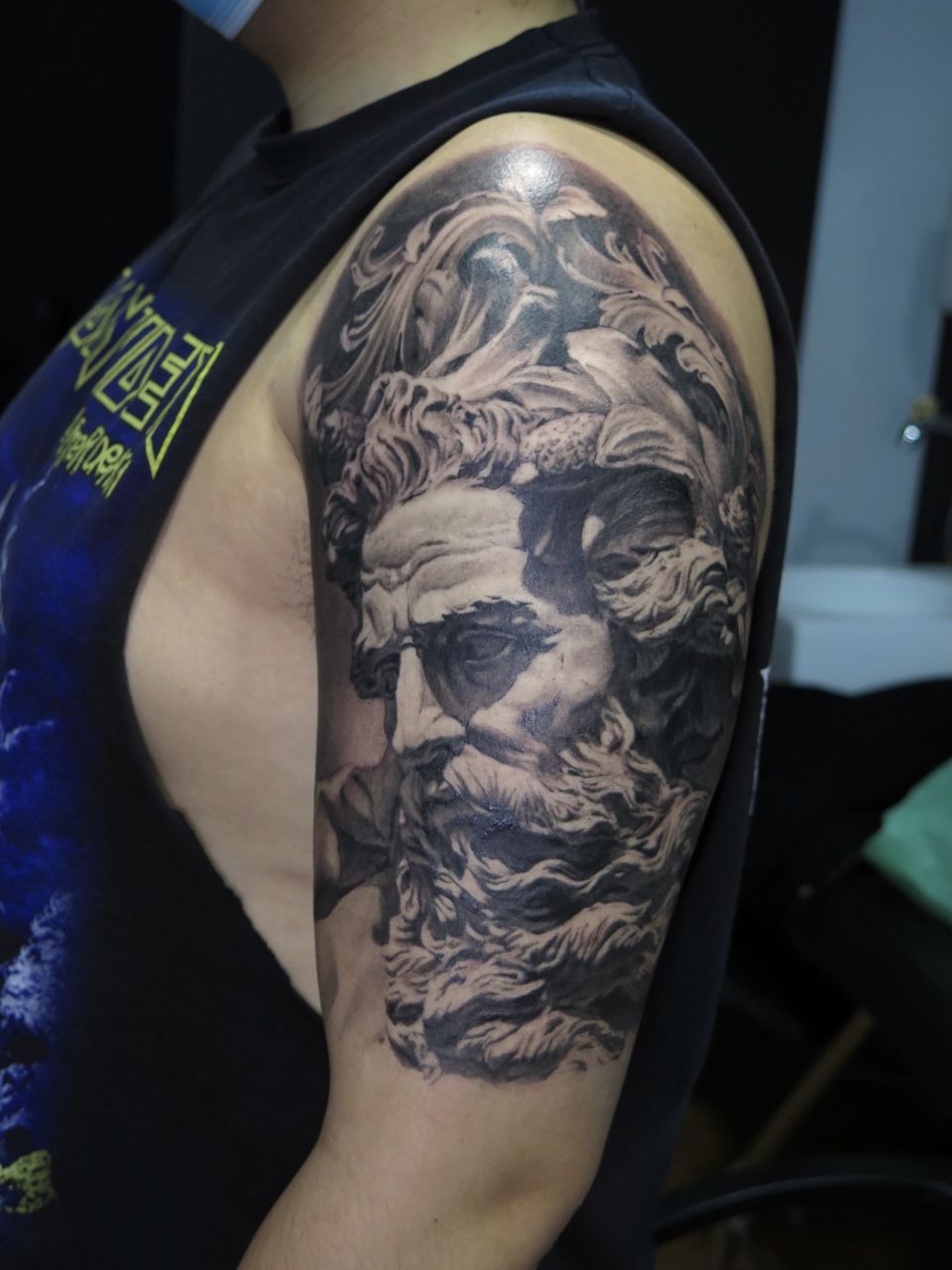 Tatuaje realista en el hombro y el brazo: busto de un dios griego barbudo al estilo de una escultura clásica, entre olas, en negro y grises