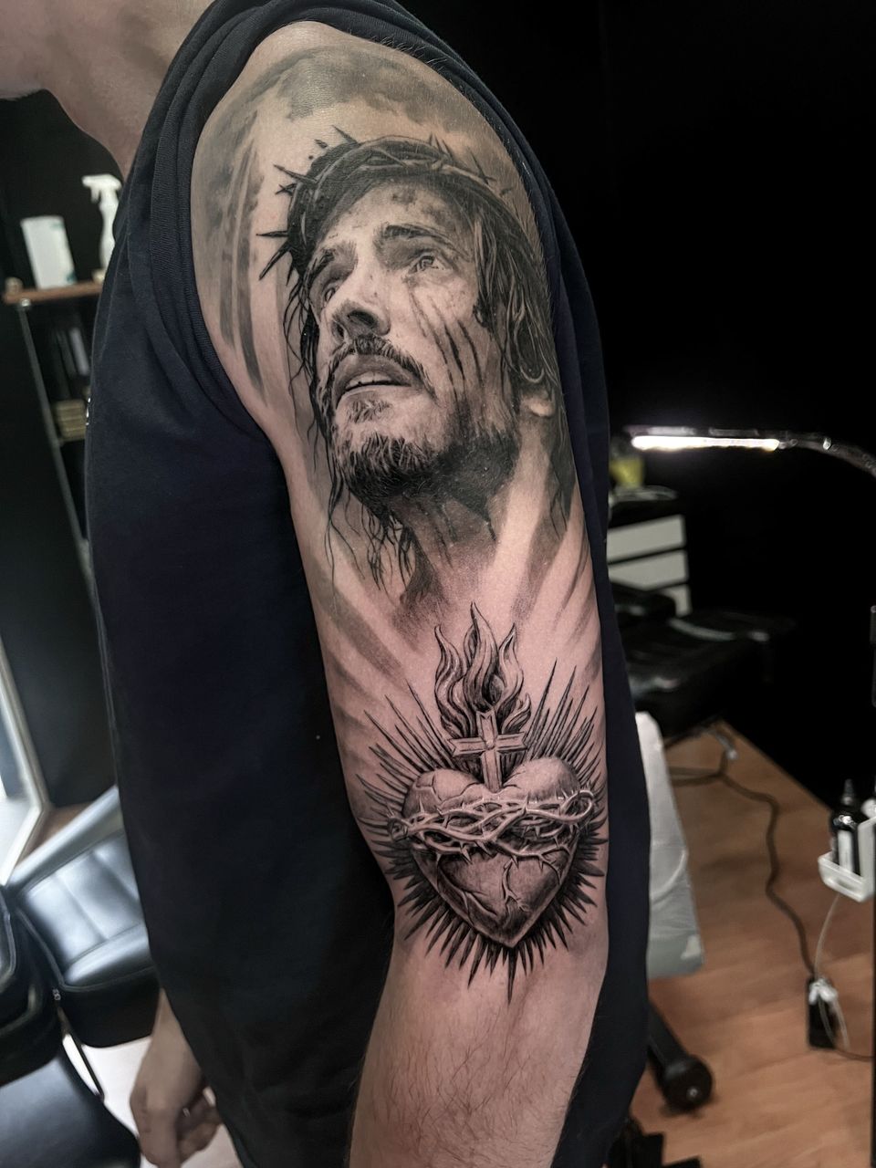 Tatuaje realista en el brazo: rostro de Jesús con corona de espinas sobre un Sagrado Corazón en llamas, en negro y grises