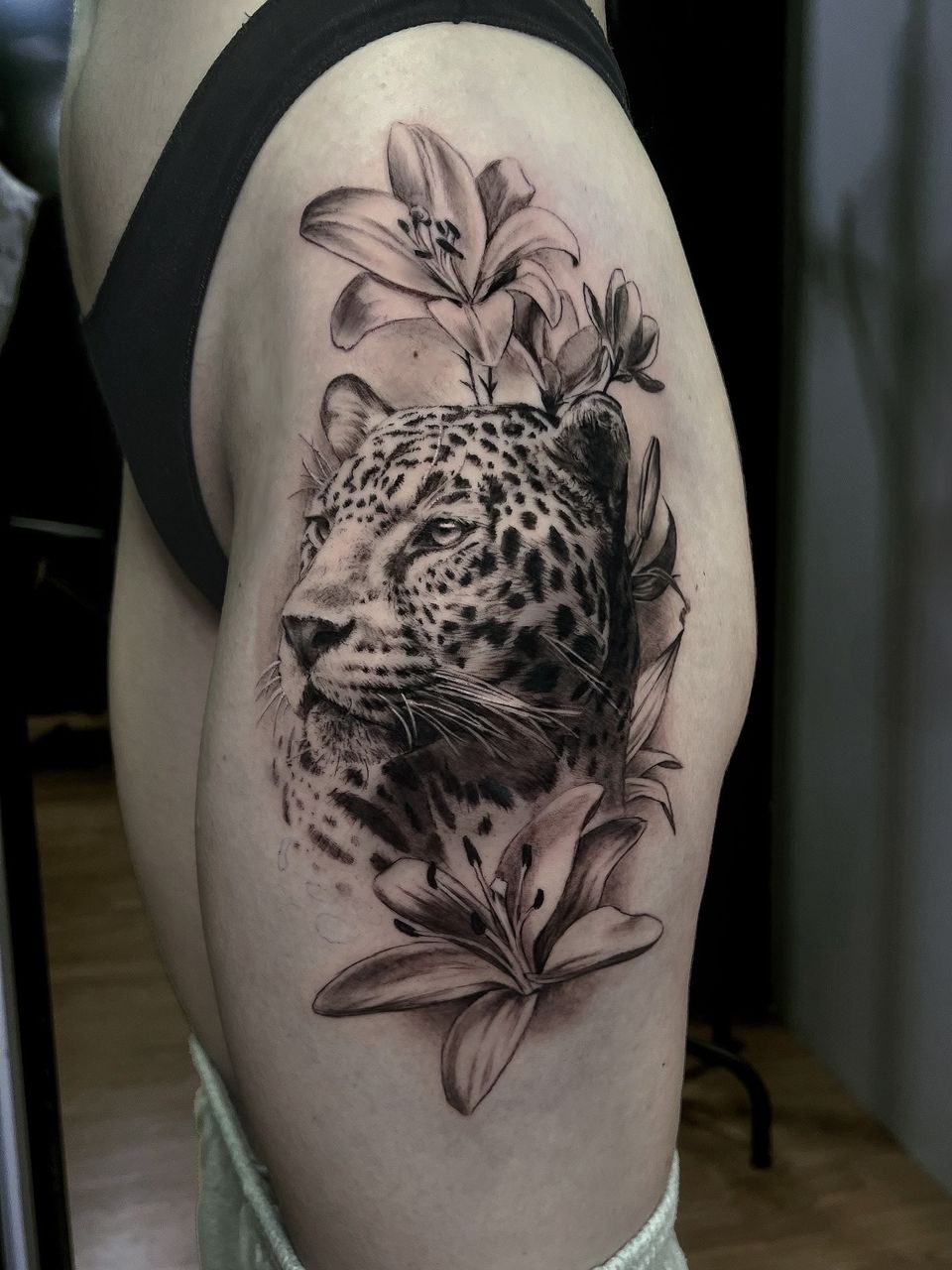 Tatuaje realista en el muslo: cabeza de leopardo entre lirios, en negro y grises