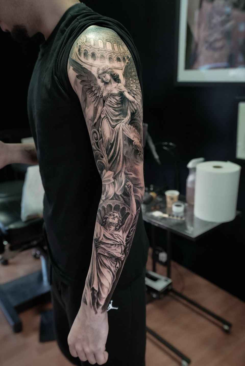 Manga completa realista en el brazo: ángel de piedra con una paloma, arcos de un coliseo y una escultura clásica mirando al cielo, en negro y grises