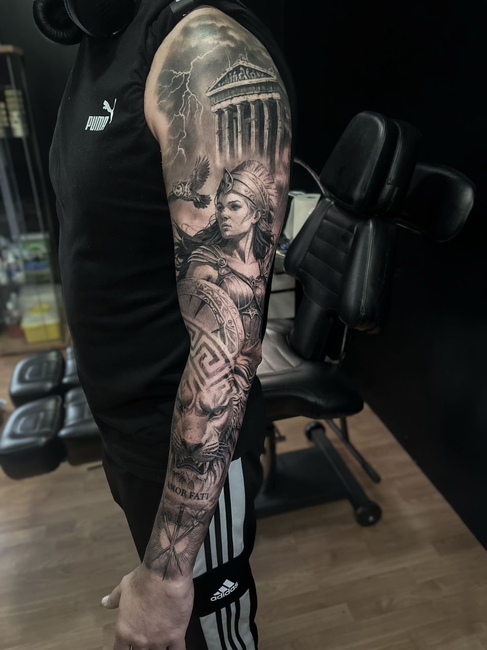 Manga completa realista en el brazo: la diosa Atenea con un búho, el Partenón bajo una tormenta, un león y la frase Amor fati, en negro y grises