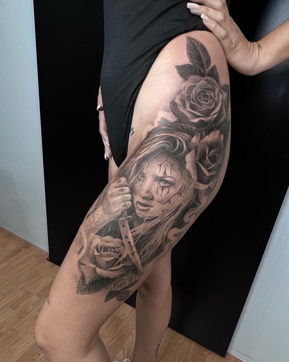 Tatuaje realista en el muslo y la cadera: mujer con maquillaje de payaso sosteniendo un cuchillo, rodeada de rosas, en negro y grises