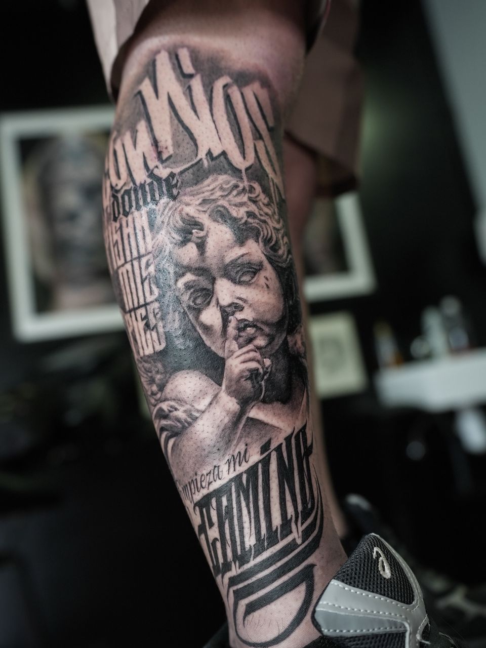 Tatuaje realista en la pierna: querubín de piedra pidiendo silencio, rodeado de una frase en letras de estilo gótico, en negro y grises