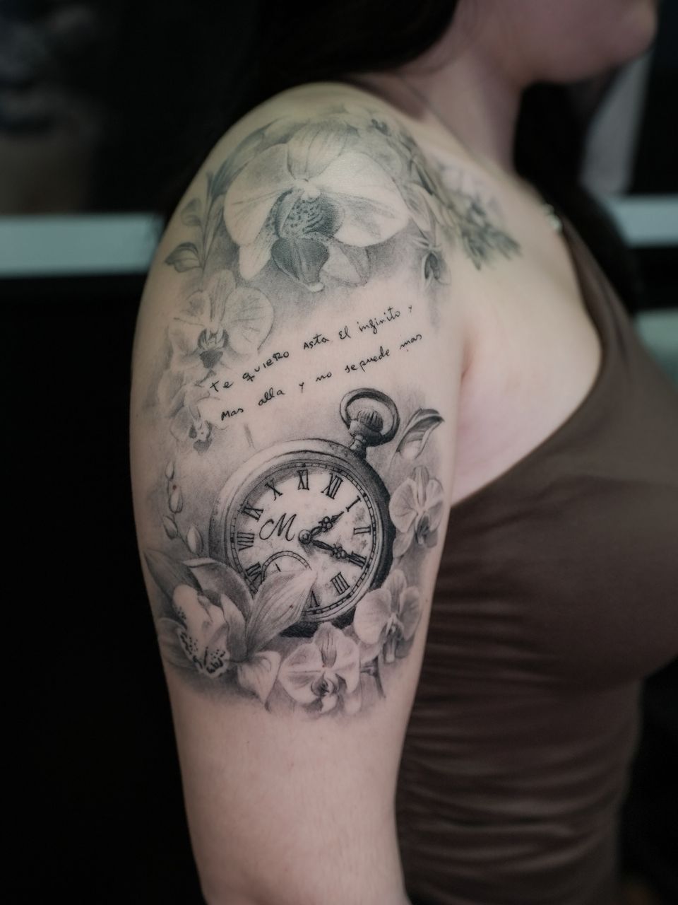 Tatuaje realista en el hombro y el brazo: reloj de bolsillo con iniciales rodeado de orquídeas y una frase manuscrita, en negro y grises