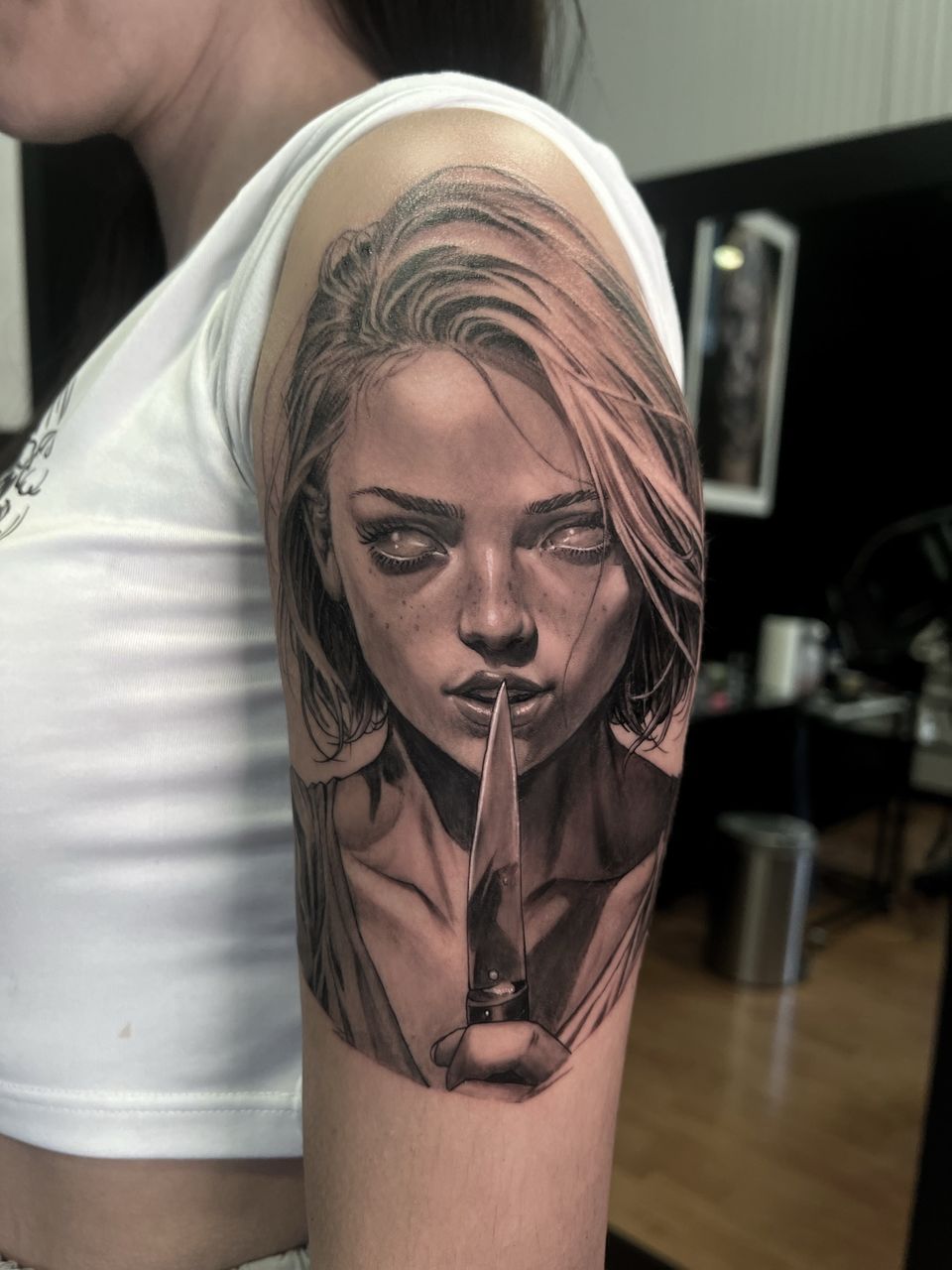 Tatuaje realista en negro y grises en la parte superior del brazo: retrato de mujer con un cuchillo frente a los labios