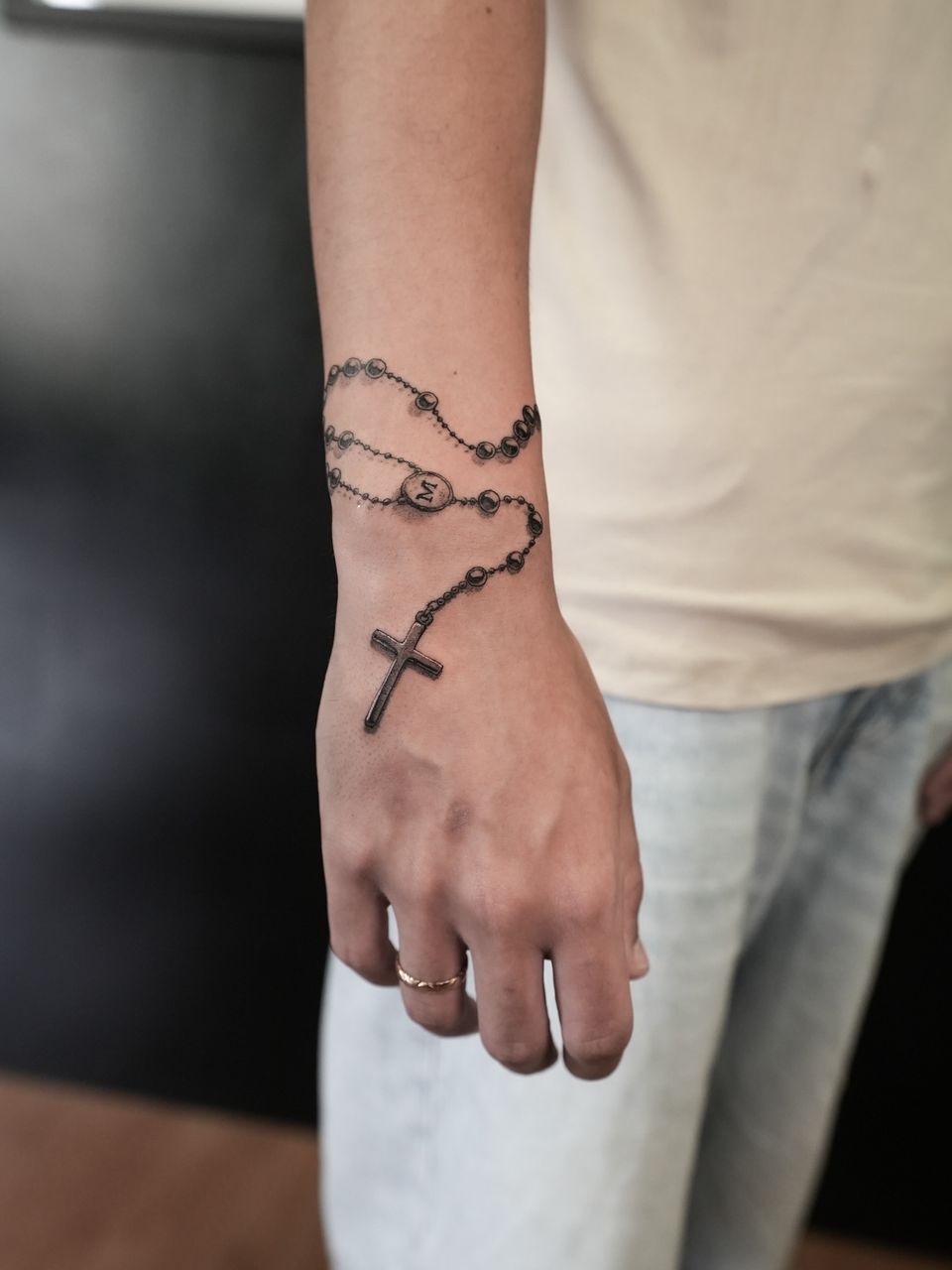 Tatuaje realista en la muñeca y el dorso de la mano: rosario enrollado con una cruz y una medalla con la letra M, en negro y grises