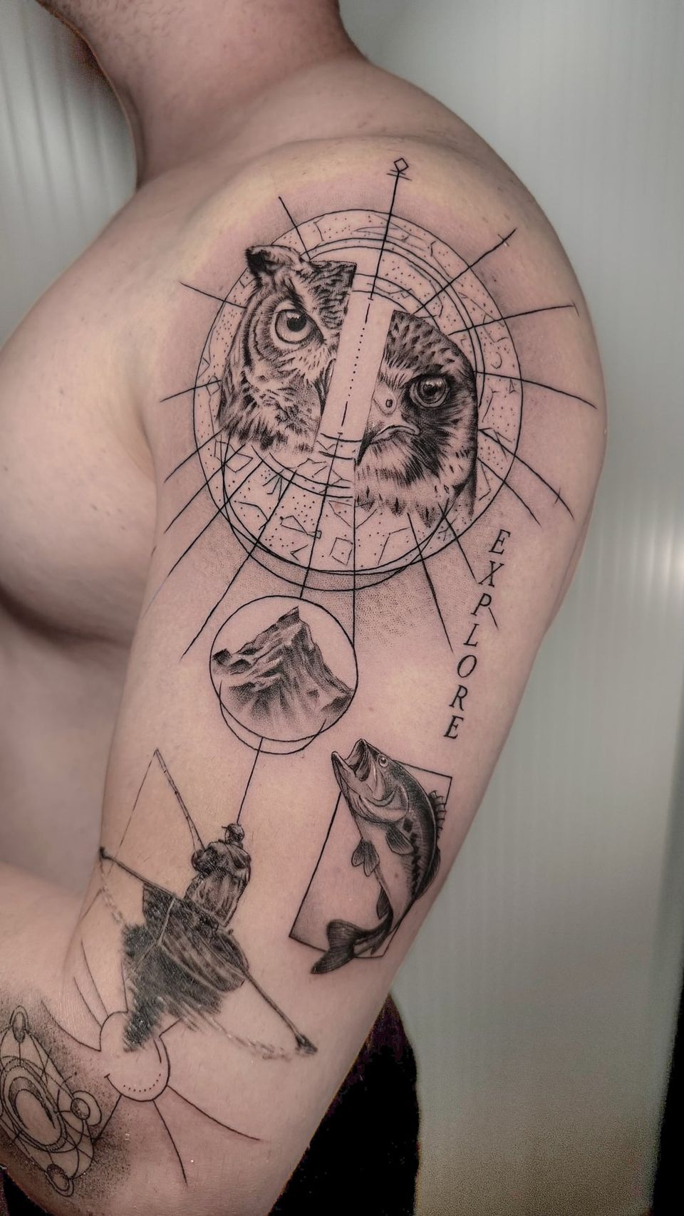 Tatuaje conceptual en el brazo: medio búho y medio halcón dentro de un círculo con constelaciones y líneas de brújula, una montaña, un pescador en su barca, una lubina saltando y la palabra «Explore»
