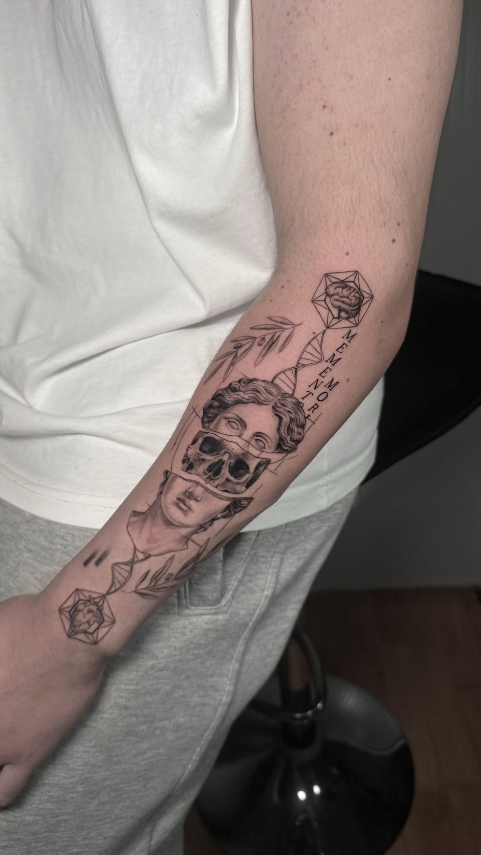 Tatuaje conceptual en el antebrazo: busto clásico partido que deja ver una calavera, una hélice de ADN que une un cerebro y un corazón dentro de poliedros, ramas de olivo y las palabras «Memento mori»