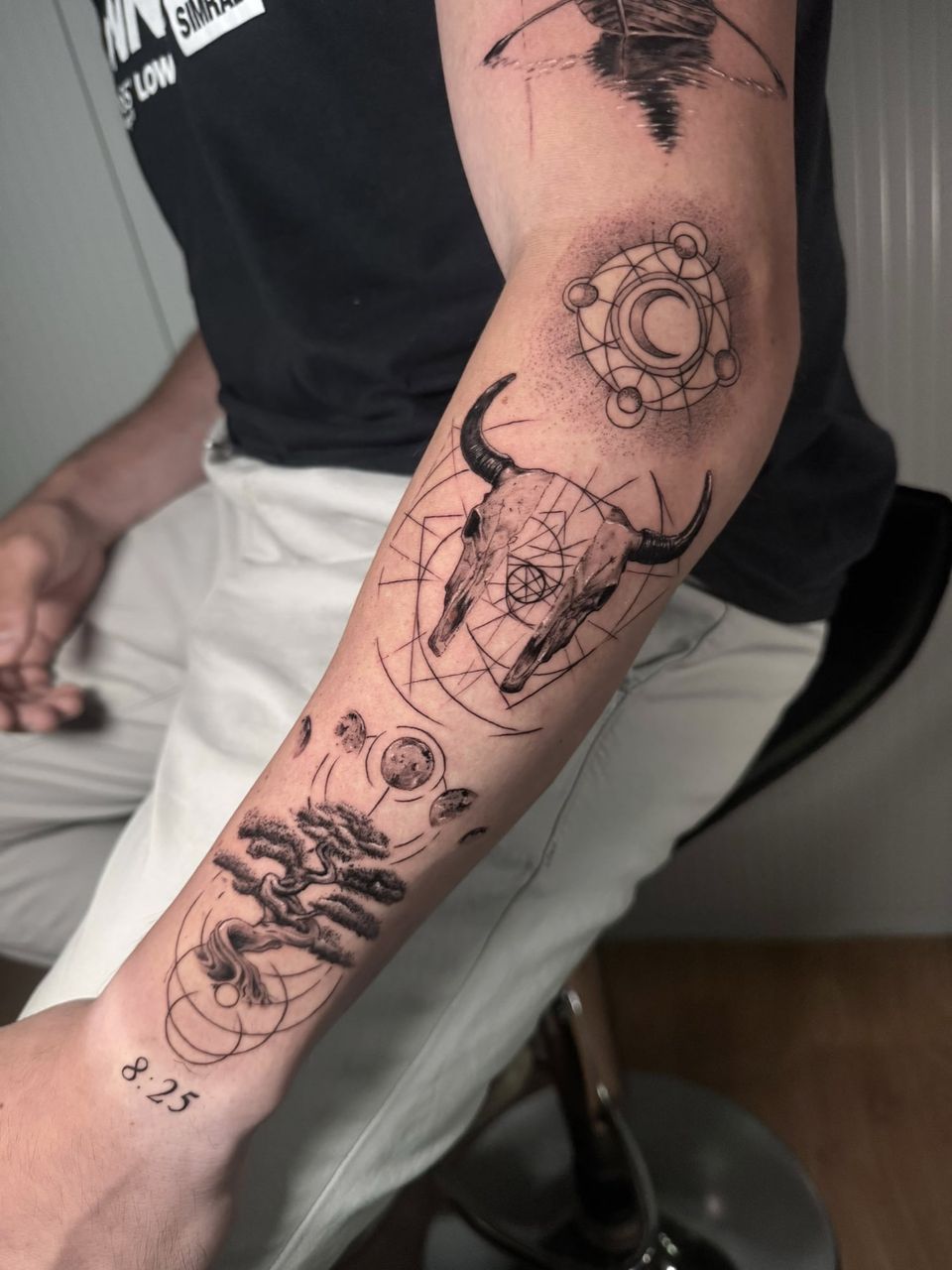 Tatuaje conceptual en el antebrazo: calavera de toro sobre líneas geométricas, un bonsái bajo las fases de la luna, un círculo con luna creciente y la hora «8:25»