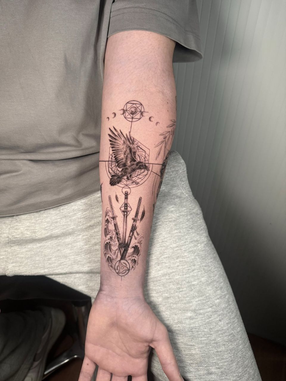 Tatuaje conceptual en el antebrazo: cuervo en vuelo sobre figuras geométricas y fases de la luna, con tres katanas entre olas