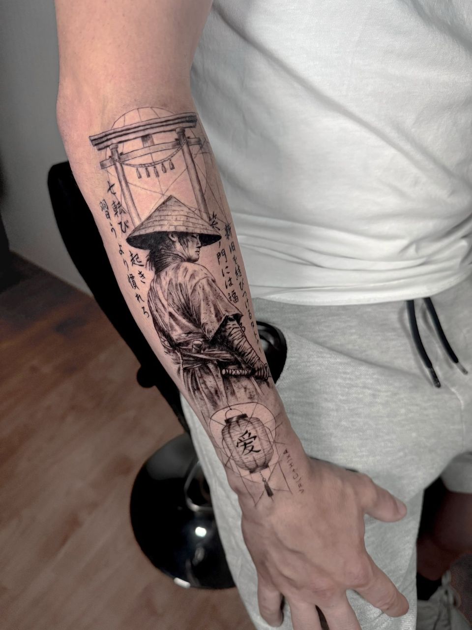 Tatuaje conceptual en el antebrazo: samurái con sombrero de paja ante una puerta torii, textos en japonés y un farolillo con el kanji del amor, sobre líneas geométricas