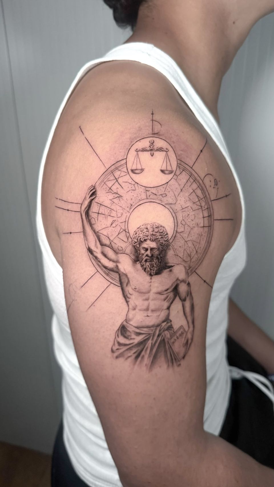 Tatuaje conceptual en el hombro: figura clásica de un titán sosteniendo una esfera celeste con constelaciones, con una balanza dentro de un círculo y líneas geométricas, en negro y grises