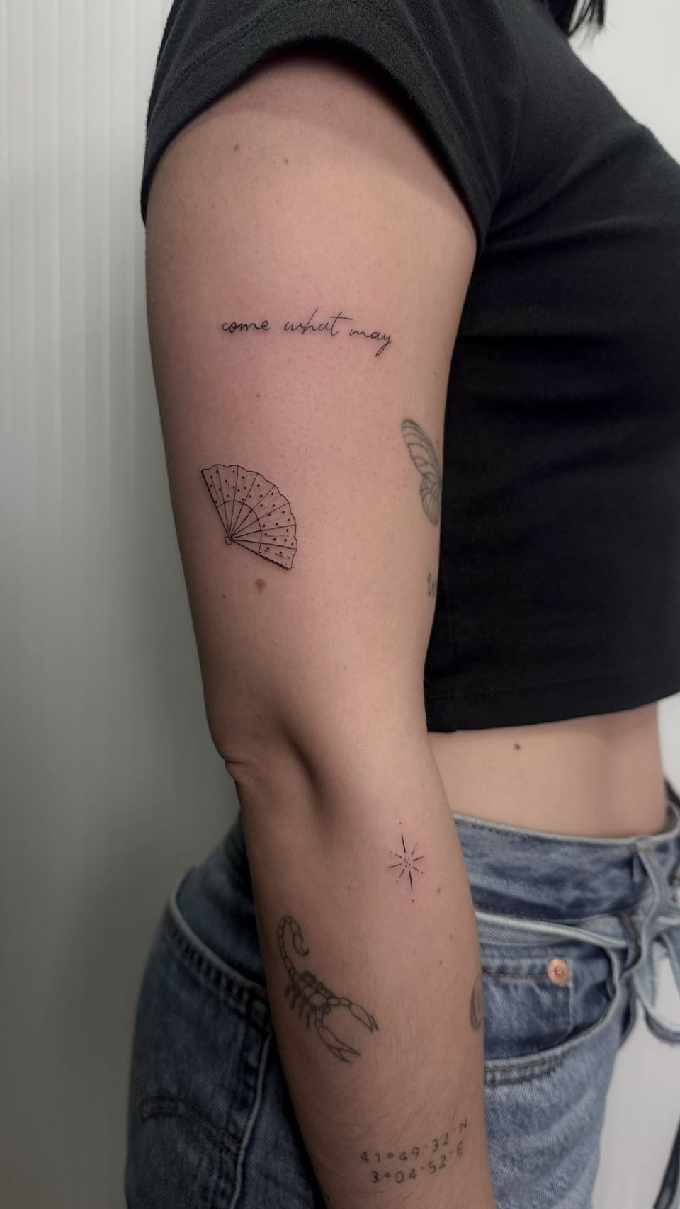 Varios tatuajes de línea fina en el brazo: la frase «come what may» en letra manuscrita, un abanico, una estrella, un escorpión y unas coordenadas