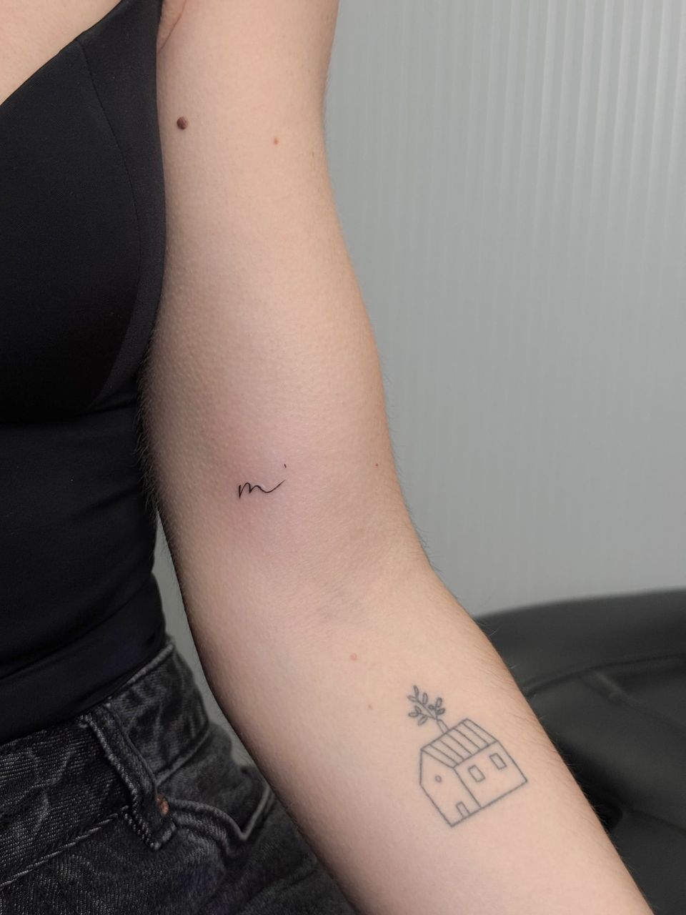 Dos tatuajes de línea fina en el brazo: una pequeña letra «m» manuscrita y una casa con una rama de olivo