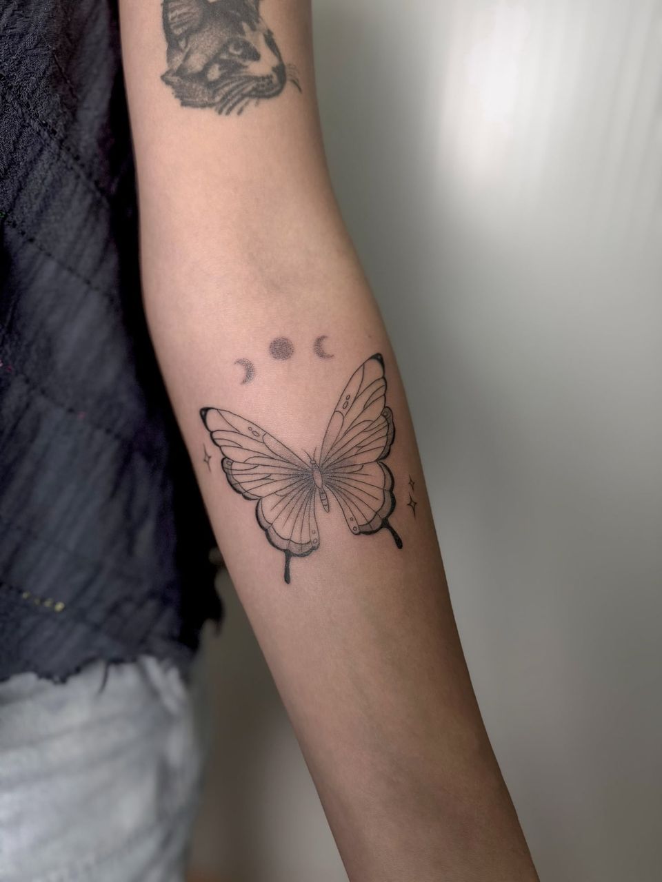 Tatuaje de línea fina en el antebrazo: mariposa con sombreado de puntos bajo tres fases de la luna y pequeñas estrellas